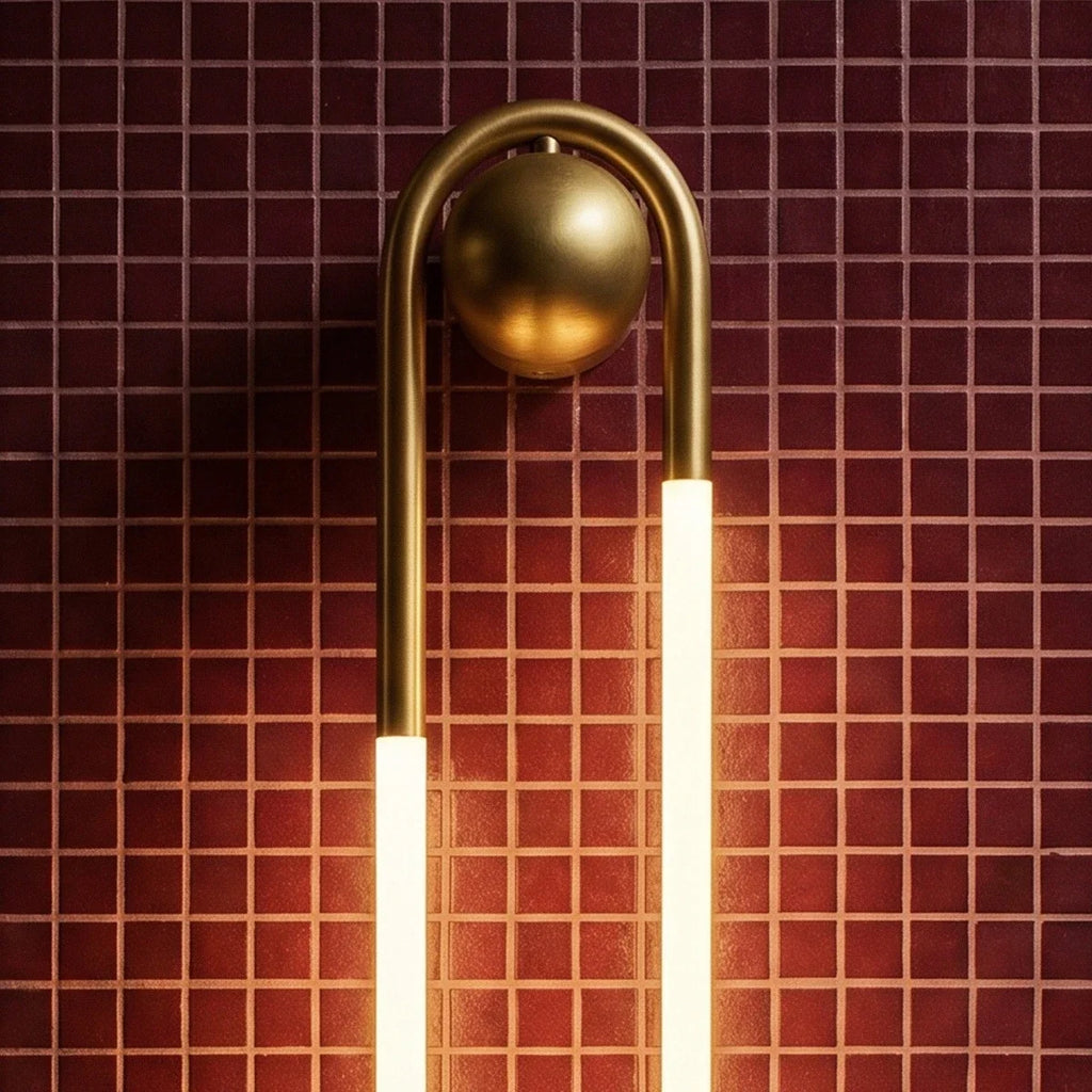 Close view of the Clérachic wall light's brushed brass arch and round ball mount above two glowing frosted tubes on red tile.