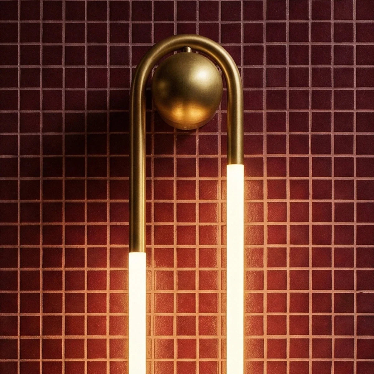 Close view of the Clérachic wall light's brushed brass arch and round ball mount above two glowing frosted tubes on red tile.