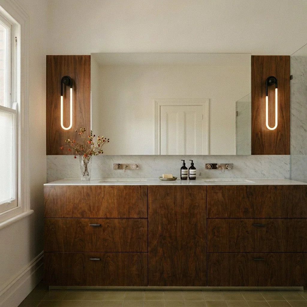 Two Clérachic black U-shaped wall lights glowing on walnut panels either side of a vanity mirror above a marble splashback.