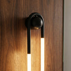 Close view of the Clérachic wall light's matt black arch and ball mount over glowing frosted tubes on a walnut wood panel.