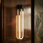 Clérachic matt black U-shaped wall light lit warm on a glossy dark wood panel, its tube reflected softly in the polished surface.