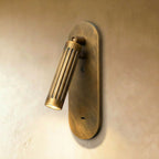 Clérachic fluted brass wall light with a tilted cylindrical head, lit warm on a slim oval backplate against plaster