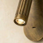 Close view of the Clérachic wall light head, a lit LED behind a fluted aged-brass cylinder, oval plate below