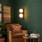 A lit Clérachic wall light with two glowing globes sits on a dark green wall above a tan armchair, a woven hanging and a small round table.