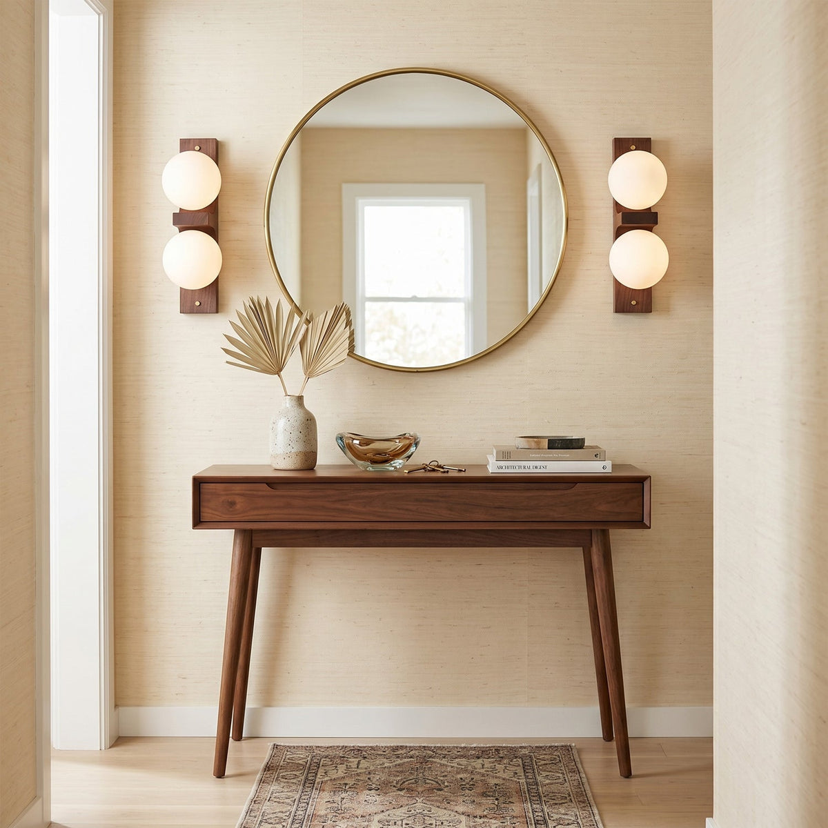 Two compact Clérachic wall lights with twin glowing globes flank a round brass-framed mirror above a slim wooden console table.