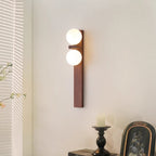 A tall Clérachic wall light with a walnut-coloured wood bar and two lit globes hangs near a window blind, with a candle and a framed picture below.