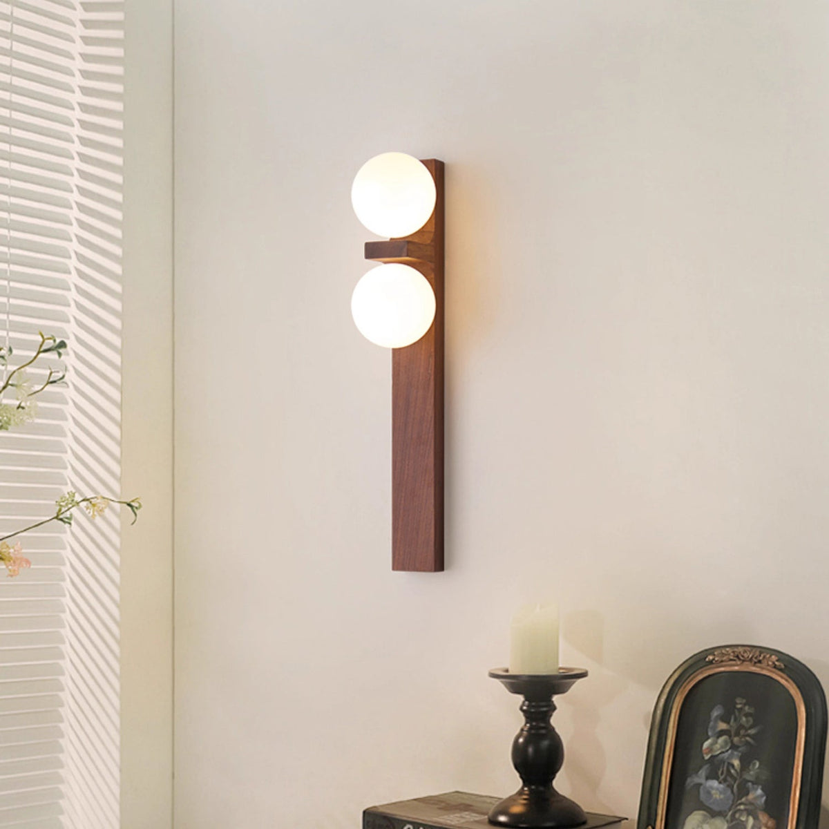 A tall Clérachic wall light with a walnut-coloured wood bar and two lit globes hangs near a window blind, with a candle and a framed picture below.