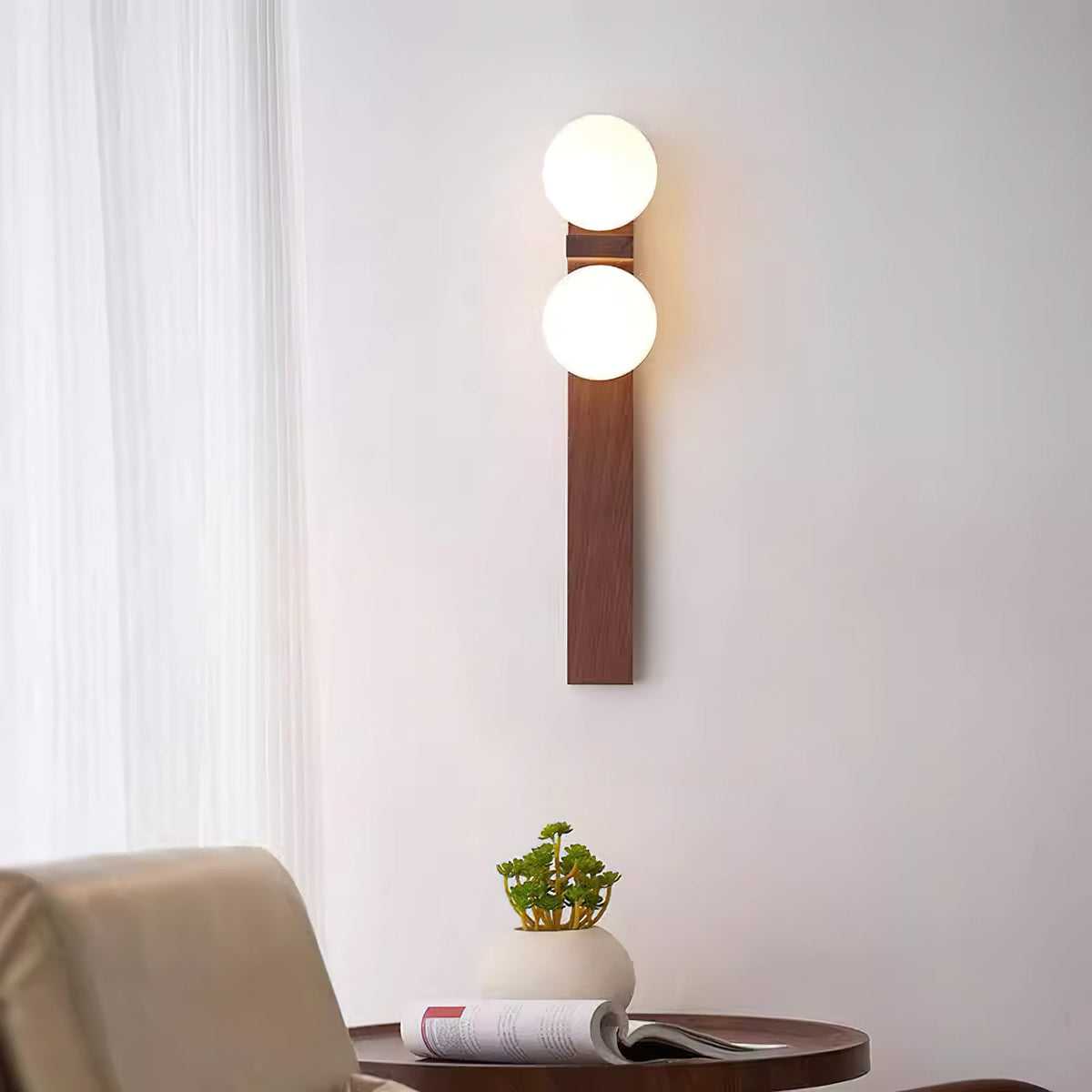 A tall Clérachic wall light with two glowing white globes hangs beside a sheer curtain, above a small round table with a potted plant.