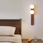 Above a wooden bedside table with a glass of water and two lemons, a tall Clérachic wall light glows beside a walnut-coloured headboard.