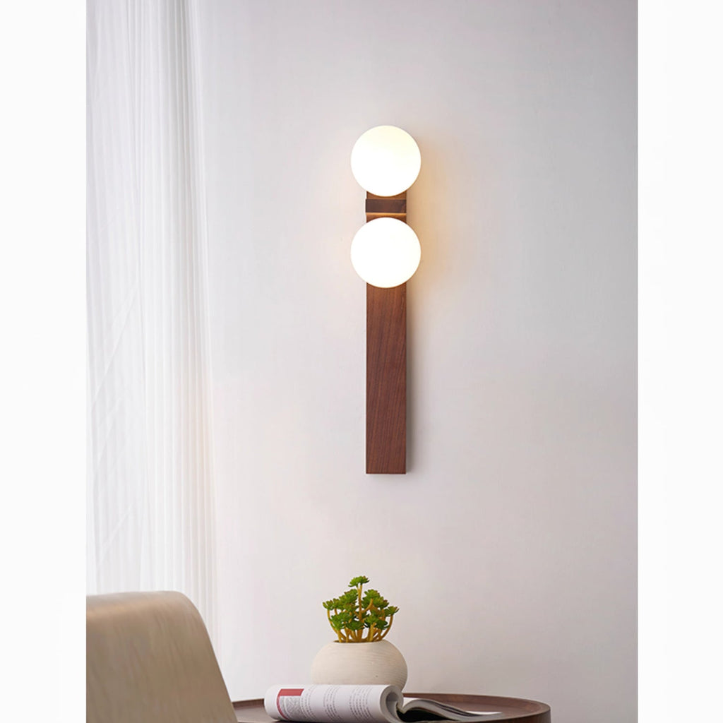 A tall Clérachic wall light with two lit globes on a long wooden bar hangs by a sheer curtain, above a table with a magazine and a plant.