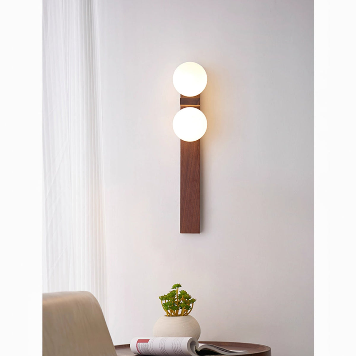 A tall Clérachic wall light with two lit globes on a long wooden bar hangs by a sheer curtain, above a table with a magazine and a plant.