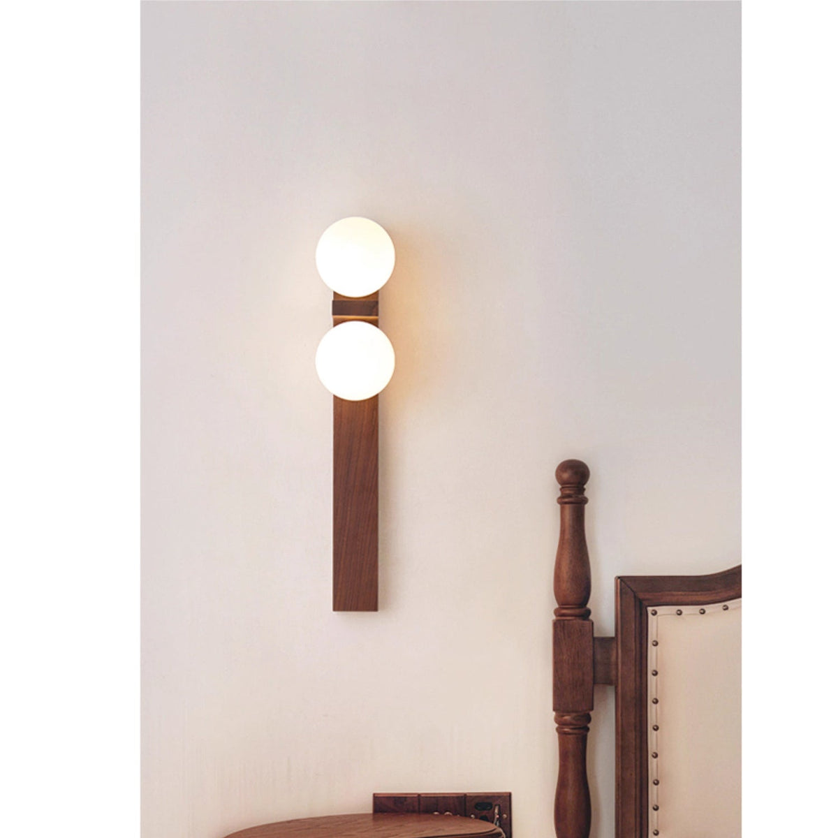 A slim Clérachic wall light with two lit globes sits high on a pale wall, above the carved post of a wooden chair.