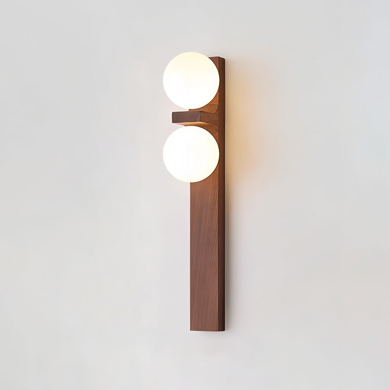 A tall Clérachic wall light with a narrow wooden bar and two lit globes casts a gentle glow on a plain pale wall.