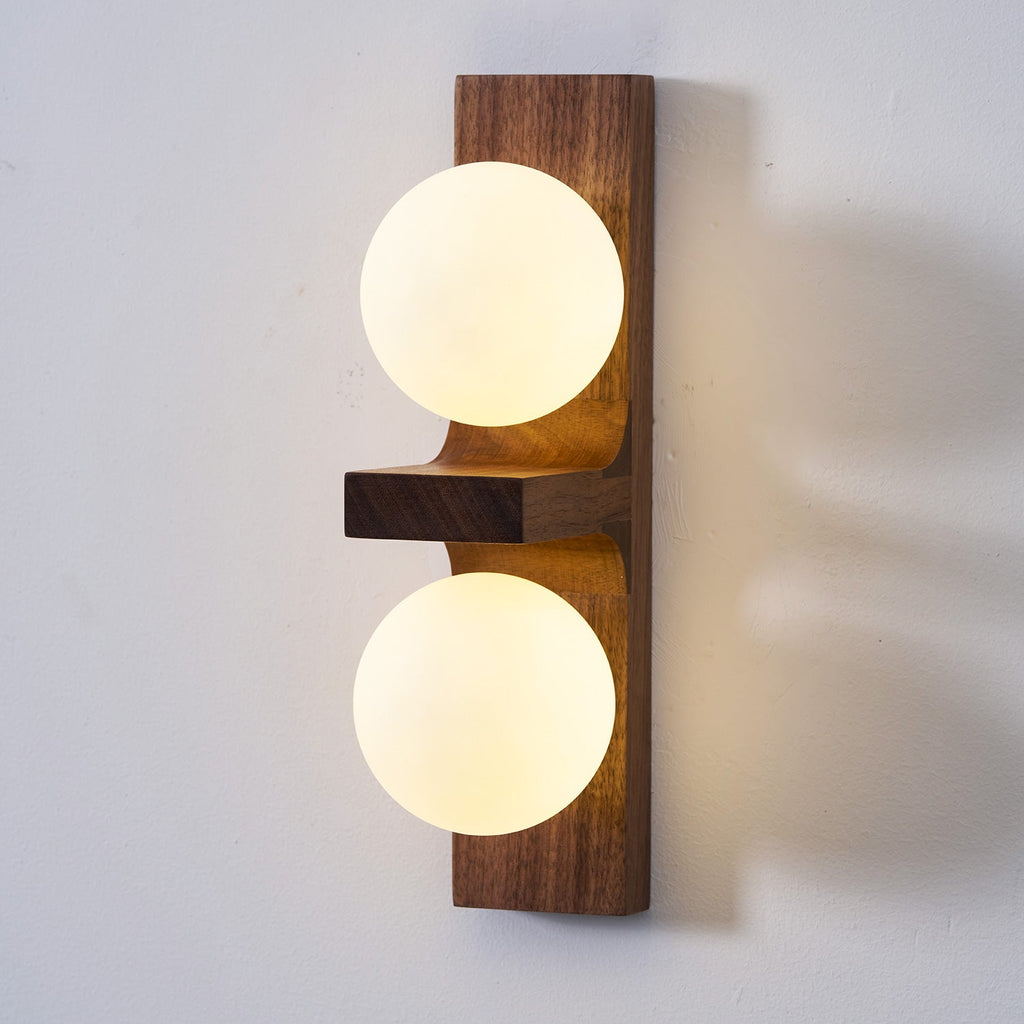 Close view of a lit Clérachic wall light, with two frosted globes on a walnut-coloured plate and a small wooden shelf between them.