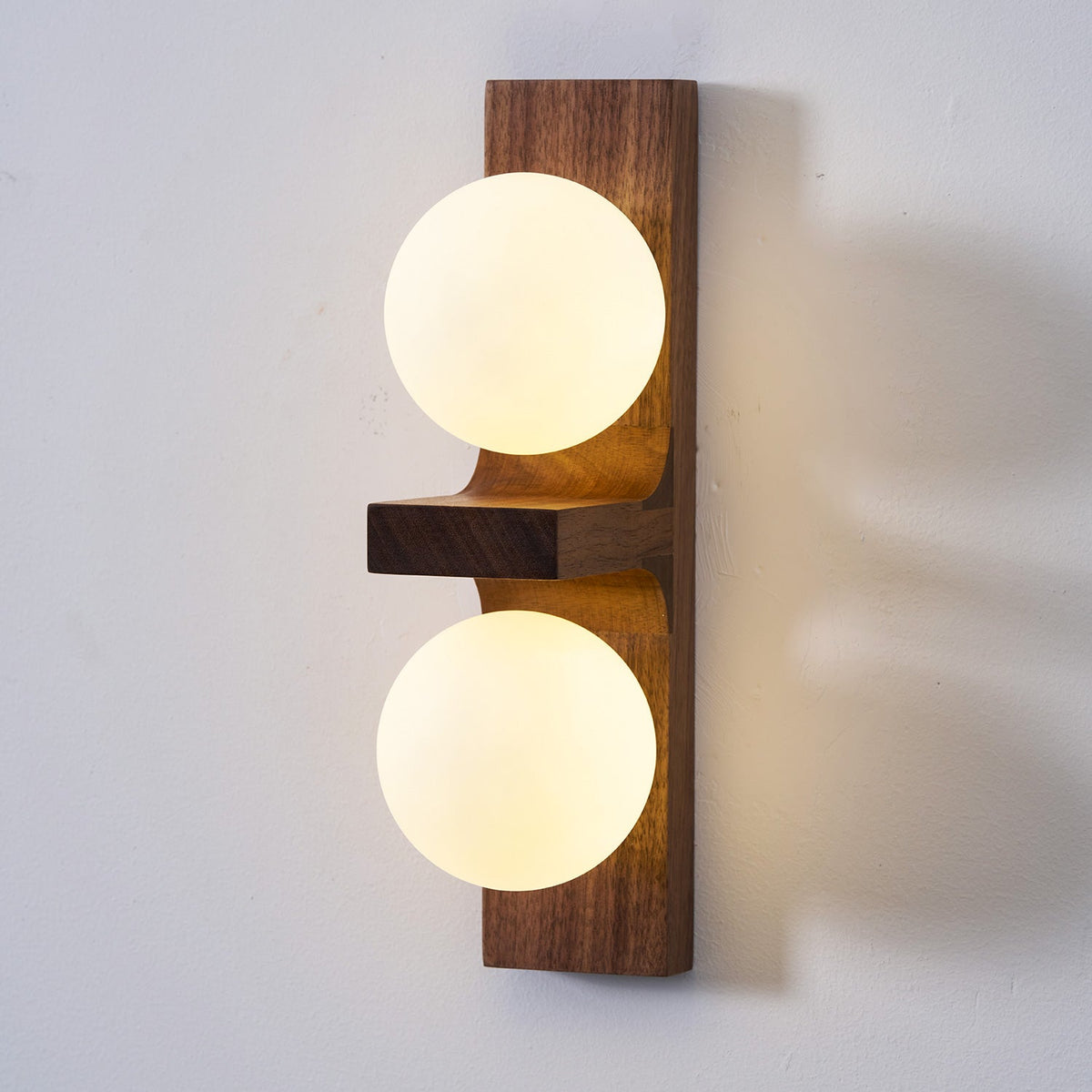 Close view of a lit Clérachic wall light, with two frosted globes on a walnut-coloured plate and a small wooden shelf between them.
