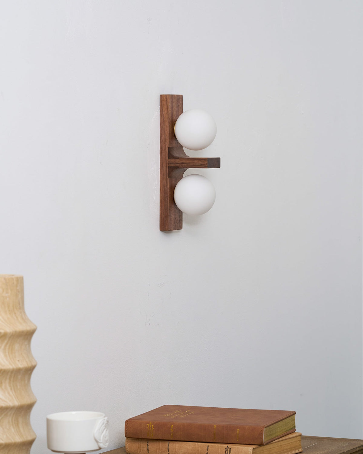 A compact Clérachic wall light with two small globes on a walnut-coloured plate hangs above a table with books and a ribbed wooden vase.