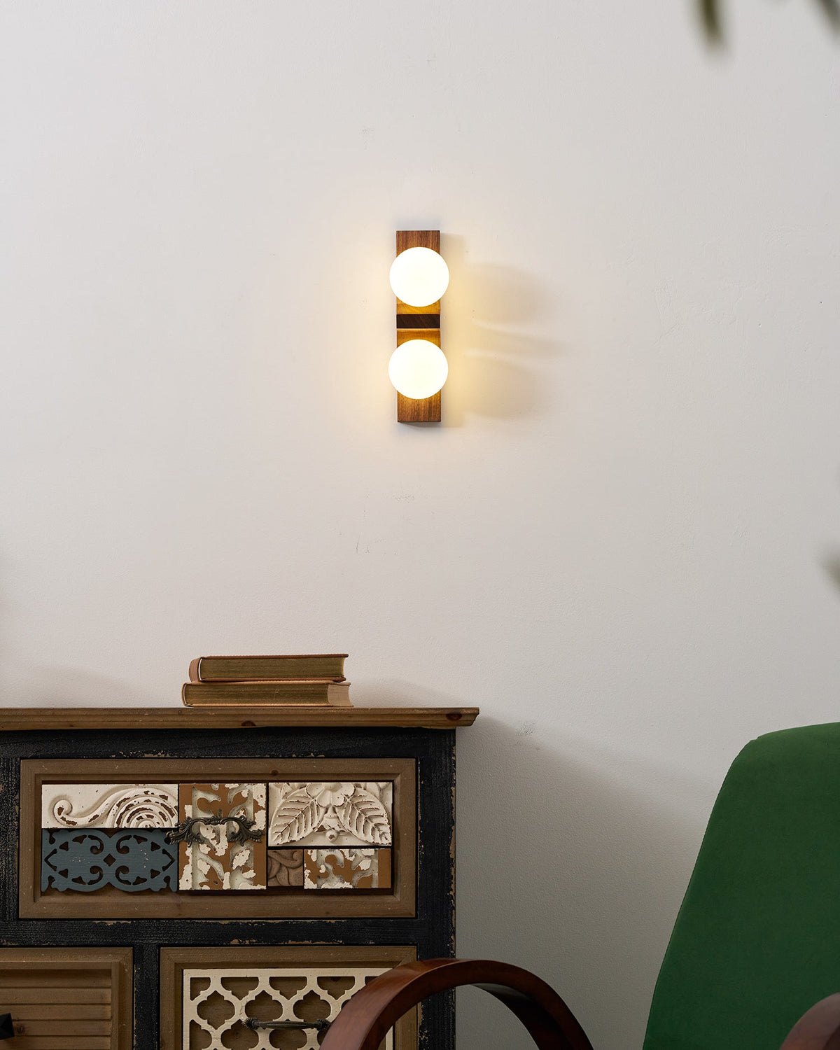 A small Clérachic wall light glows on a white wall above a painted wooden cabinet with a stack of books, beside a green armchair.