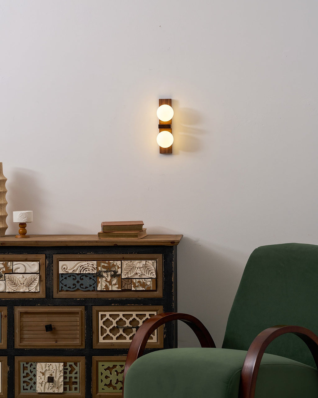 A lit Clérachic wall light on a white wall above a patterned wooden cabinet, with a green armchair in the corner.