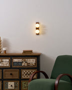 A lit Clérachic wall light on a white wall above a patterned wooden cabinet, with a green armchair in the corner.
