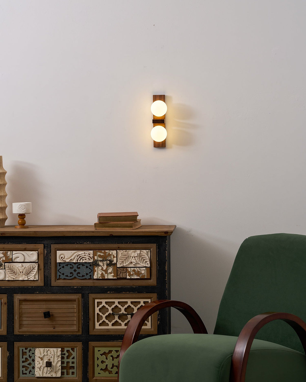 A lit Clérachic wall light on a white wall above a patterned wooden cabinet, with a green armchair in the corner.