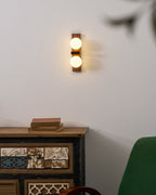 A Clérachic wall light with two warmly lit globes hangs above a carved wooden cabinet with books, near the edge of a green chair.