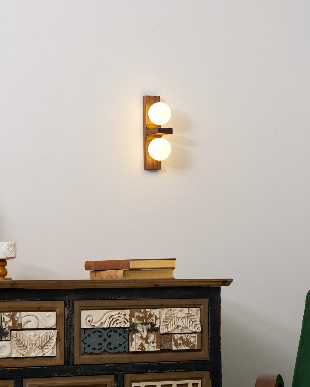 A Clérachic wall light with two glowing globes on a walnut-coloured plate hangs above a patterned cabinet with a stack of old books.