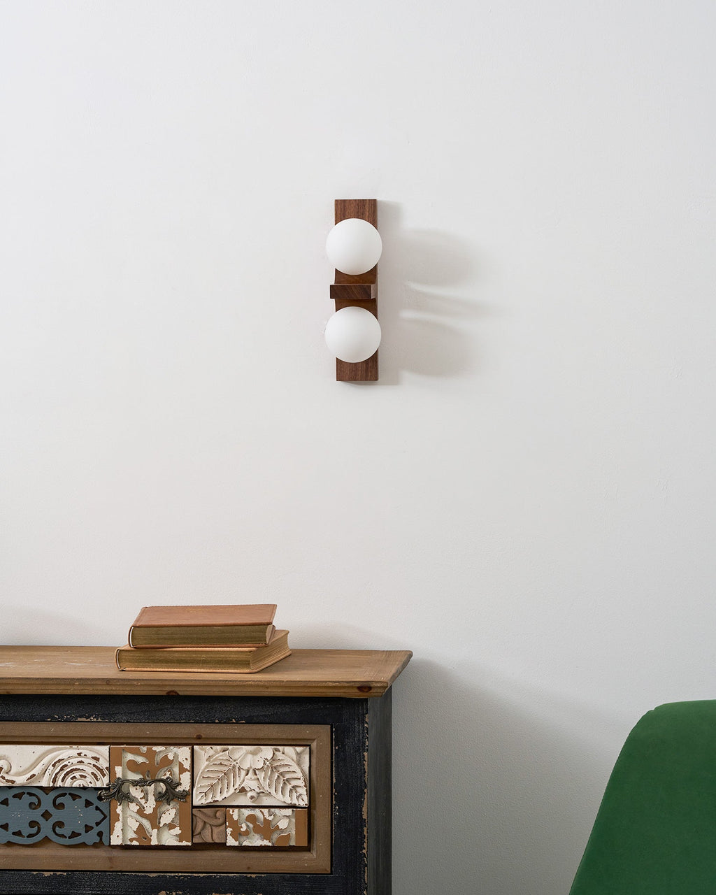 An unlit Clérachic wall light with two white globes hangs above a cabinet topped with books, beside a green armchair.