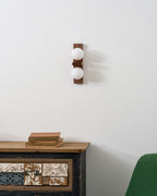 An unlit Clérachic wall light with two white globes hangs above a cabinet topped with books, beside a green armchair.