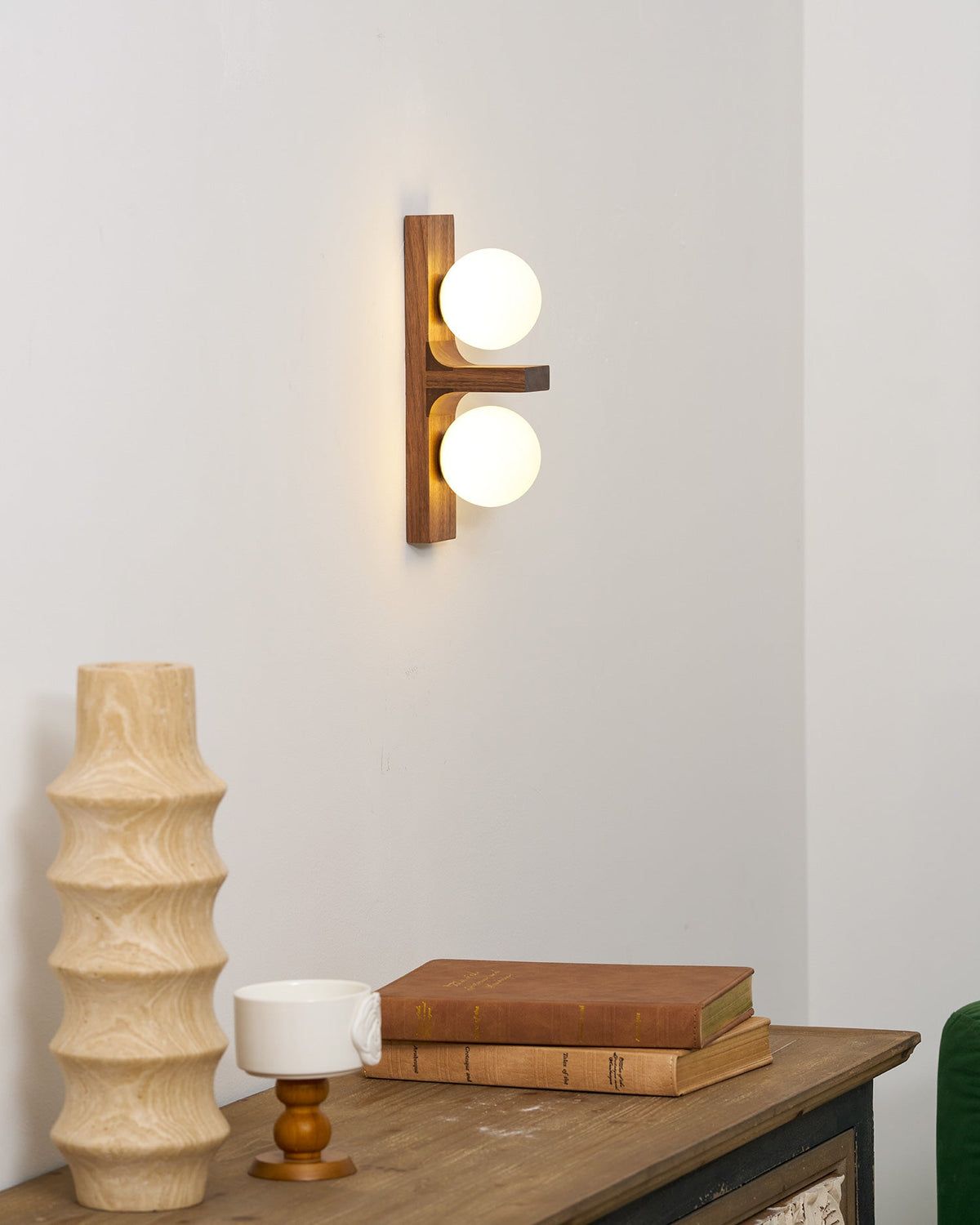 A lit Clérachic wall light hangs above a wooden table with books, a pale carved vase and a small cup, in a white-walled corner.