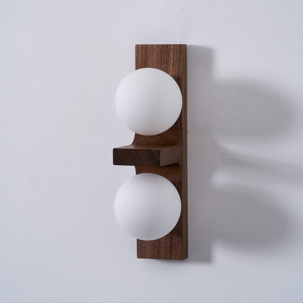 Side view of a Clérachic wall light with a walnut-coloured plate, a flat wooden shelf and two matt white globes, casting a soft shadow.