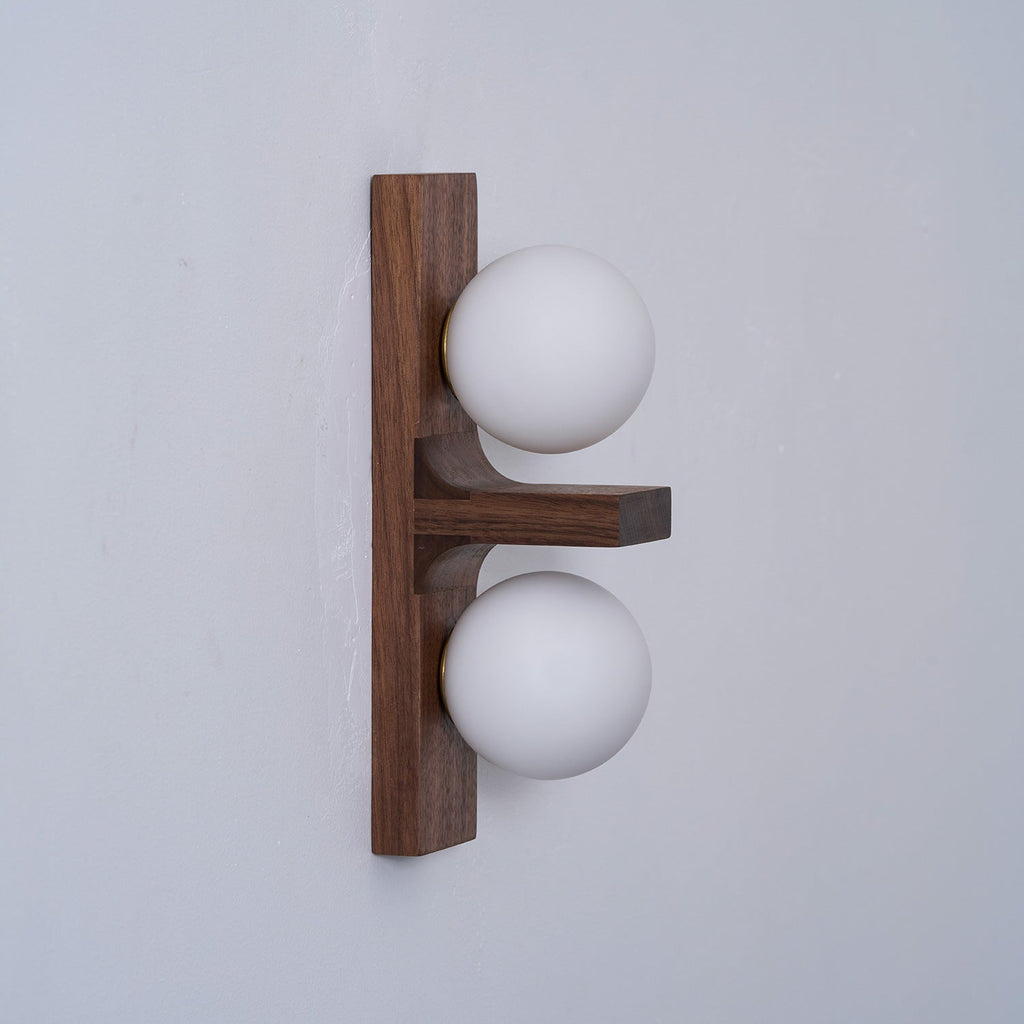 Seen from the side, a Clérachic wall light shows the grain of its walnut-coloured wood plate, a small shelf and two frosted globes.