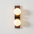 A Clérachic wall light with two bright frosted globes and a darker walnut-coloured plate casts a warm glow on a white wall.