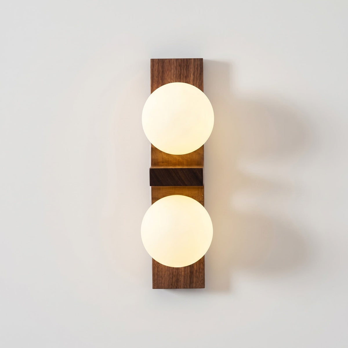 A Clérachic wall light with two bright frosted globes and a darker walnut-coloured plate casts a warm glow on a white wall.