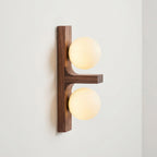 A lit Clérachic wall light with two warm-white globes on a walnut-coloured wood plate, seen at an angle against a pale wall.