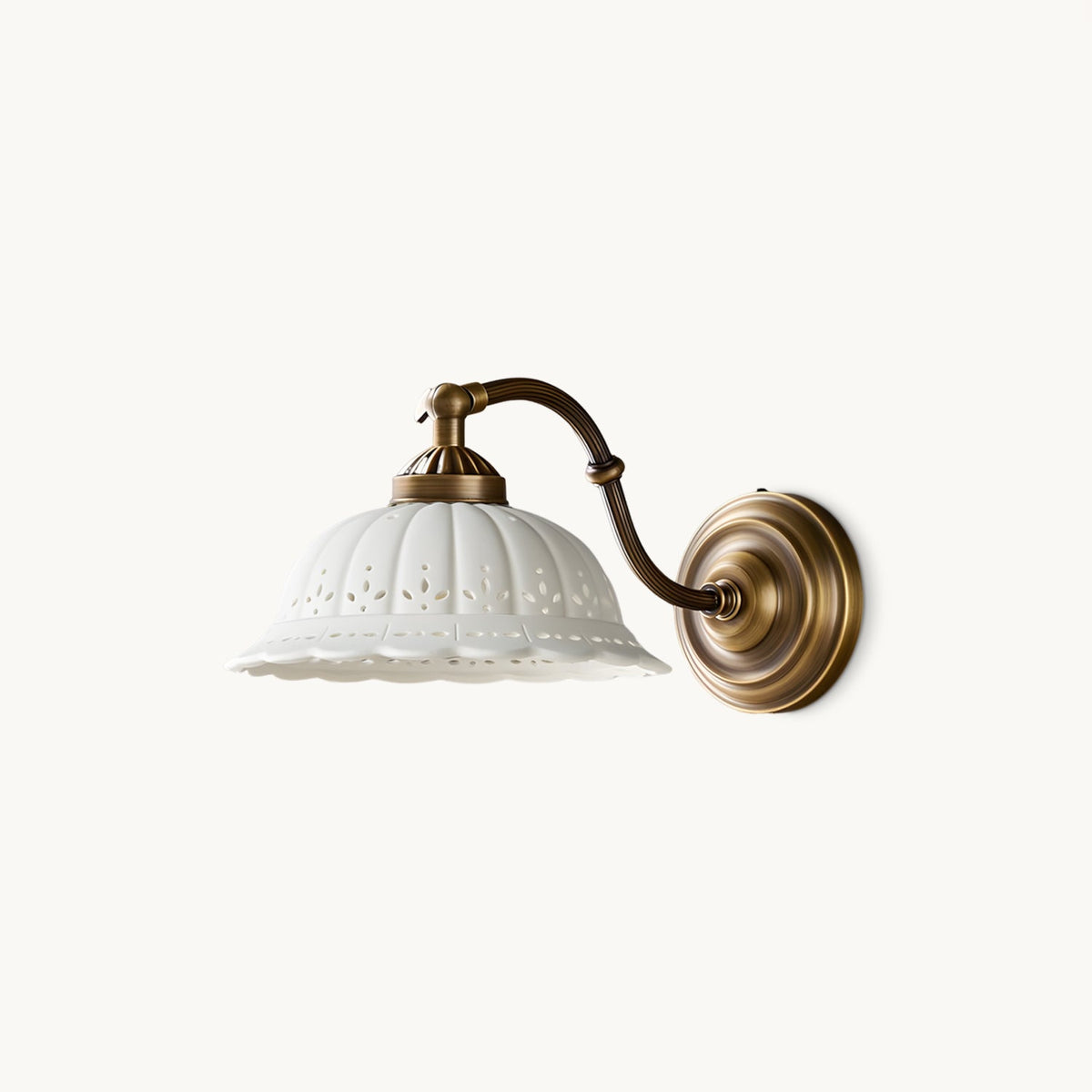 On a plain white backdrop, a Clérachic wall light with a white ceramic shade pierced with small holes, on a curved antique brass arm and round backplate.