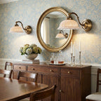 Two Clérachic wall lights with pierced white ceramic shades hang either side of a round gilt mirror, above a wooden sideboard with flowers.