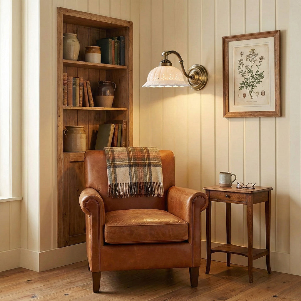 A lit Clérachic wall light with a white ceramic shade and brass arm glows above a leather armchair, beside a wooden bookcase.