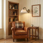 A lit Clérachic wall light with a white ceramic shade and brass arm glows above a leather armchair, beside a wooden bookcase.