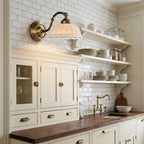 Over open shelves and a white tiled kitchen wall, a Clérachic wall light with a pierced white ceramic shade on an antique brass arm.