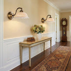 Two lit Clérachic wall lights with pierced ceramic shades hang above a console table in a hallway with white panelling and a runner rug.