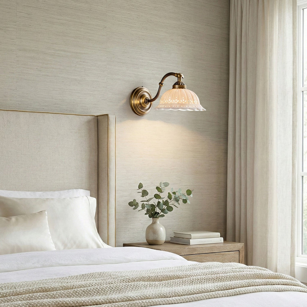 A Clérachic wall light with a scalloped white ceramic shade glows above a bed with a linen headboard and a small vase on the bedside table.