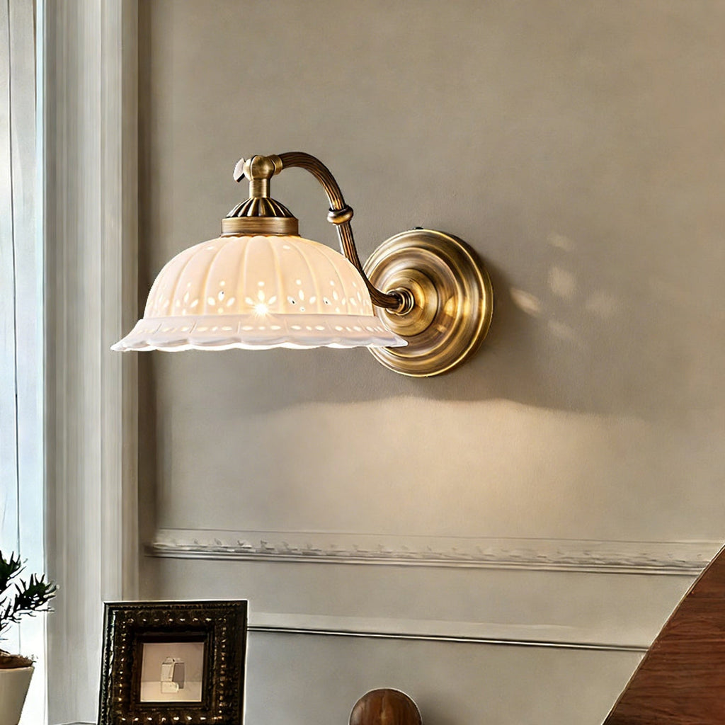 Close view of a Clérachic wall light, with a scalloped white ceramic shade and a round antique brass backplate, lit softly against a grey wall.