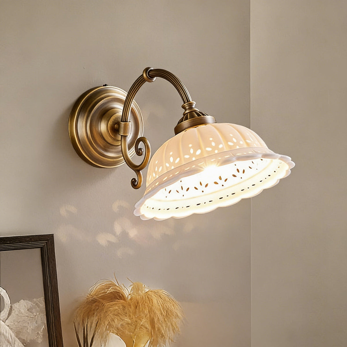 Close view of a lit Clérachic wall light, with a flared ceramic shade pierced with tiny holes, hung from a scrolled brass arm.