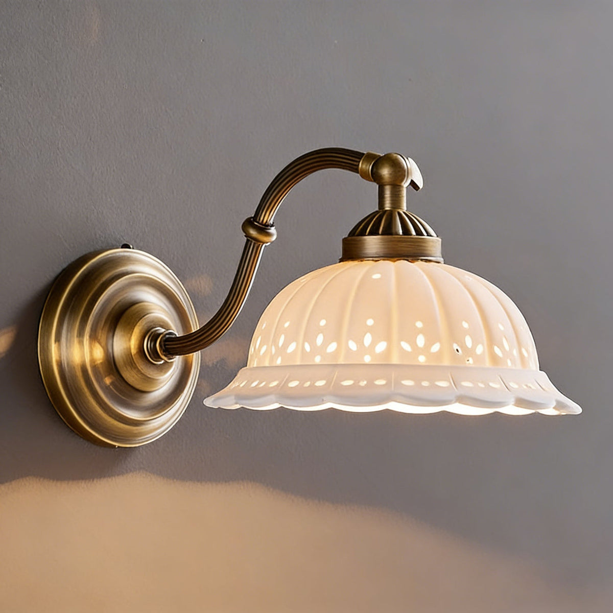 Warm light pools on a grey wall from a Clérachic wall light with a curved brass arm and a ribbed white ceramic shade.