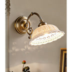 Seen from slightly below, a lit Clérachic wall light shows the pierced pattern around the rim of its white ceramic shade, with a scrolled brass arm.