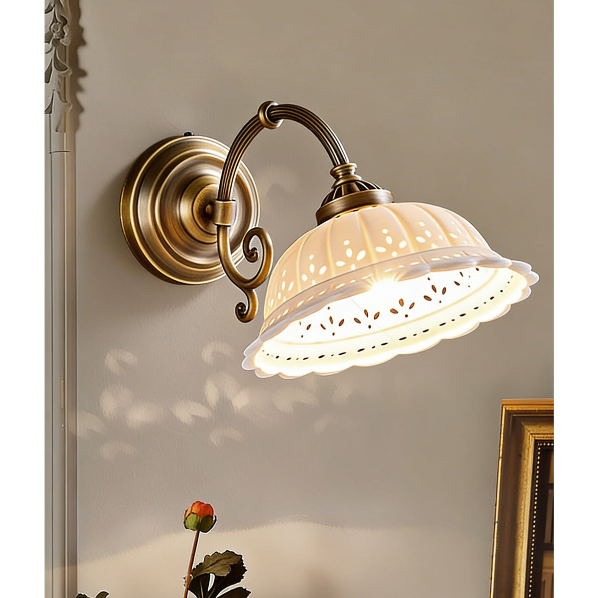 Seen from slightly below, a lit Clérachic wall light shows the pierced pattern around the rim of its white ceramic shade, with a scrolled brass arm.