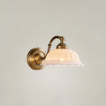 A lit Clérachic wall light with a rounded brass backplate, curved arm and ribbed white ceramic shade, against a plain pale wall.