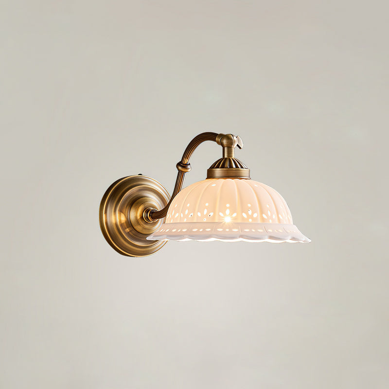 A lit Clérachic wall light with a rounded brass backplate, curved arm and ribbed white ceramic shade, against a plain pale wall.