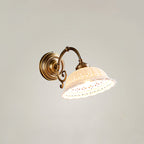 A lit Clérachic wall light with a scrolled brass arm and a flared, pierced ceramic shade, set against a plain pale wall.