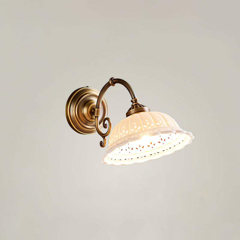 A lit Clérachic wall light with a scrolled brass arm and a flared, pierced ceramic shade, set against a plain pale wall.